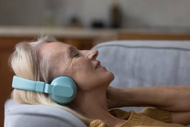 Kapalı yüzlü, yaşlı, 50 'li yaşlarda kaygısız bir kadın kanepeye uzanıp kablosuz kulaklıkla favori müziğin tadını çıkarıyor. Çevrimiçi dijital yayın hizmetleri başvuru kullanımı, hobi, ev kavramında stressiz zaman geçirme