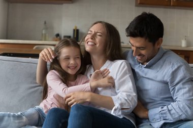 Genç, çekici ebeveynler ve anaokulu sevimli kızı kanepede oturuyor, gülüyorlar, çok eğleniyorlar, birbirlerini gıdıklıyorlar ve evde eğleniyorlar.