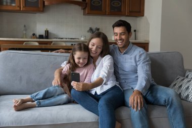 Anaokulu kızı olan bir çift kanepeye oturup eğleniyor, video seyir defteri izliyor, fotoğraf çekiyor, akrabalarıyla uzaktan konuşmanın tadını çıkarıyor ve akıllı telefon kullanıyor. Kablosuz modern teknoloji konseptiyle aile eğlencesi