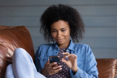 Akıllı telefon kullanan, evdeki rahat koltukta dinlenen, telefon ekranına bakan, sosyal ağdaki arkadaşlarla sohbet eden, alışveriş yapan, video izleyen, boş zamanlardan zevk alan Afrikalı Amerikalı bir kadın.