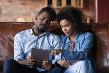 Afro-Amerikalı çift tablet kullanıyor, internetten alışveriş yapıyor, evdeki koltukta oturuyor, gülümseyen kadın ekranı işaret ediyor, internet mağazasından mal seçiyor, sosyal ağda mesaj okuyor.