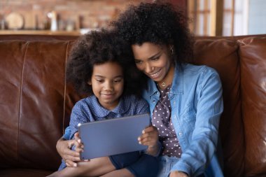Kapalı kapılar ardında gülümseyen Afrikalı Amerikalı anne ve küçük kızı tablet kullanıyor, evdeki koltukta dinleniyor, mutlu genç anne ve 7 'lerin kızı modern cihazlarla vakit geçiriyorlar.