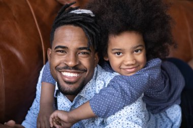 Kafadan çekilen portre gülümseyen Afrikalı Amerikalı baba küçük kızı sırtında taşıyor, evdeki rahat kanepede dinleniyor, sevimli 7 'lerin kız çocuğu genç babaya sarılıyor, aile birlikte vakit geçiriyor.