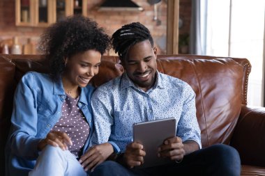 Mutlu Afro-Amerikan çift tablet kullanıyor, kanepede birlikte oturuyor, gülümsüyor kadın ve erkek cihaz ekranına bakıyor, komik videolar izliyor, boş zamanların tadını çıkarıyor, sohbet ediyor veya online alışveriş yapıyor.