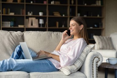 Mutlu meşgul genç kadın kanepede dinleniyor, dizüstü bilgisayar kullanıyor, cep telefonuyla konuşuyor. Serbest çalışan, evden çalışan uzaktan çalışan, cep telefonunda danışmanlık yapan. Uzak iş, çoklu görev