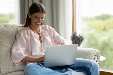 Neşeli genç ev sahibi kadın dizüstü bilgisayar kullanıyor, daktilo ediyor, internette sohbet ediyor, internette geziniyor, film izliyor, rahat bir kanepeden alışveriş yapıyor. Serbest çalışan, evden çalışan uzaktan çalışan.