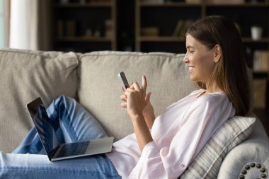 Mutlu neşeli kız çeşitli dijital cihazlar kullanmaktan hoşlanıyor, evdeki koltukta dinleniyor, dizüstü bilgisayarı tutuyor, cep telefonuyla sohbet ediyor, uygulamadan kısa mesaj atıyor. İnternet bağımlılığı