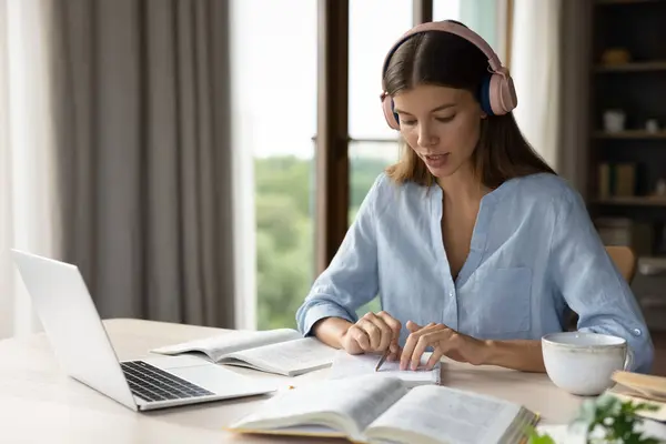 stock image Focused engaged student girl in big headphones studying foreign language, listening audio lesson at laptop, reading notes out loud, doing exercises from open book, school, college homework task