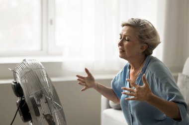 Yaşlı kadın kendini sıcak hissediyor, vantilatörün önünde oturuyor, vantilatörle serinlemeye çalışıyor, oturma odasındaki klimasız aşırı sıcaktan muzdarip, terli ve kurumuş görünüyor.