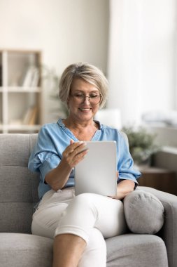 Gülümseyen olgun bir kadın evde tablet kullanarak gözlük takıyor, haber okuyor, aile veya arkadaşlarla mesajlar paylaşıyor, bilgiye ve çevrimiçi eğlenceye erişiyor. Modern teknolojiyi kullanarak sosyal bağlantı