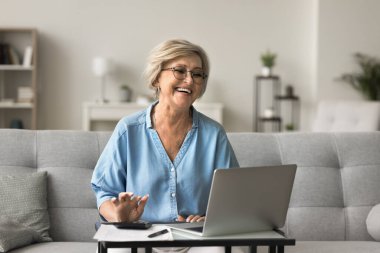 Gülümseyen yaşlı kadın, dizüstü bilgisayar, hesap makinesi ve kağıtlarla aynı masaya oturur, ev faturalarını veya vergilerini internetten öder, aylık giderleri ve gelirleri kontrol eder, finans yönetimi modern teknoloji kullanır