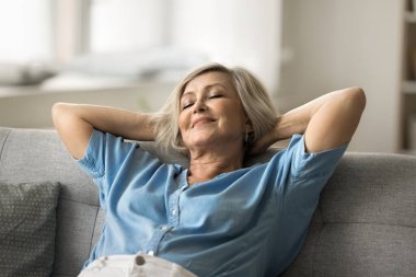 Sakin, yaşlı bir kadın kanepeye yaslanıyor, elleri başının arkasında, gözleri kapalı dinleniyor, yüzünde huzurlu ve memnun bir ifadeyle, rahat bir oturma odasında rahatlık ve rahatlığın tadını çıkarıyor.