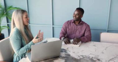 Mutlu, güzel, beyaz, profesyonel bir kız toplantı masasında Afrikalı bir erkek meslektaşla konuşuyor, laptopta konuşuyor, şirket arkadaşıyla çevrimdışı iletişimin keyfini çıkarıyor.