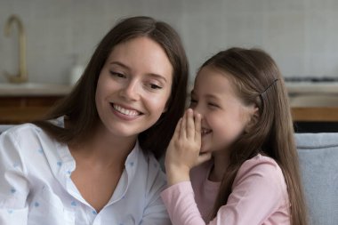 Neşeli heyecanlı küçük kız mutlu annesiyle sırrını paylaşıyor, kulağına fısıldıyor, gülüyor. Anne ve tatlı kız arkadaş olmanın keyfini çıkarıyorlar, güven, aile eğlencesi, sohbet, kanepede dinlenme.