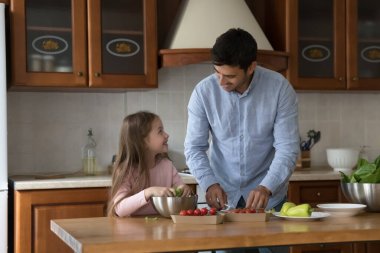 Neşeli yakışıklı baba küçük kıza salata pişirmeyi öğretiyor. Mutlu baba ve kız mutfakta birlikte yemek hazırlıyor, taze sebzeleri dilimliyor, marul yapraklarını parçalıyor, konuşuyor, gülüyor.