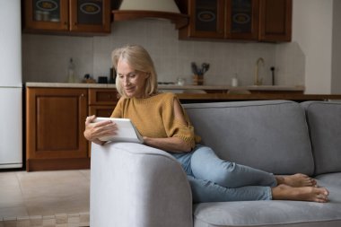 Evde tablet kullanan mutlu, olgun bir kadın, rahat bir koltukta dinleniyor, online medya içeriğini izliyor, ecommerce uygulaması ile alışveriş yapıyor. İletişim, yaşlılık, eğlence konsepti