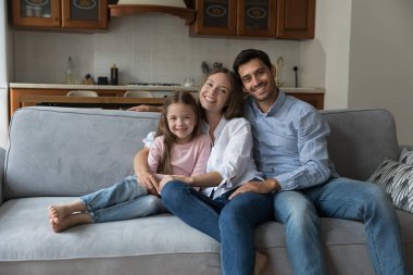 Mutlu bir çift ebeveyn ve küçük kız evdeki rahat koltukta oturmuş kameraya bakıyor ve gülümsüyor. Ebeveynlik, gayrimenkul, sigorta kavramı