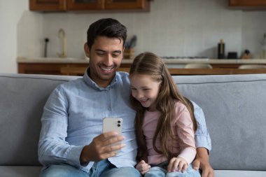 Mutlu Latin baba ve küçük beyaz kız akıllı telefondan video konferans görüşmesi yapıyorlar, internette sohbet ediyorlar, gülüyorlar, cep telefonlarını evdeki kanepede tutuyorlar, ekranlara bakıyorlar.