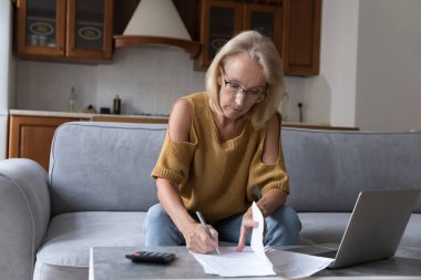 Gözlüklü ciddi bir orta yaşlı ev sahibi, yerel belgeleri gözden geçiriyor, ödeme faturaları, hesap makinesinde ve laptopta kağıtlar yazıyor, tasarrufları, vergileri, sigorta ücretlerini sayıyor.