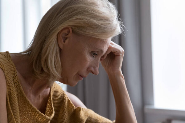 Sad thoughtful lonely elderly lady suffering from apathy, depression, mental disease, looking into vacancy, thinking over health problems, bad news, loss, grief, touching head. Side view