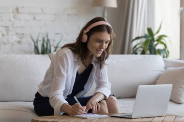 Kulaklık takan kadın kanepeye oturup e-öğreniyor, dizüstü bilgisayar kullanıyor, webinar izliyor, yeni bilgiler edinmek için güler yüzlü ve motive olmuş görünmesini sağlıyor. Çevrimiçi öğretmen kavramına göre eğitim dersi