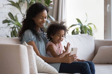 Ebeveyn kontrolü, internetteki çocuk güvenliği, yeni mobil uygulama, modern kablosuz teknoloji kullanımı kavramı. Afrikalı anne ve kız kanepede oturup cep telefonlarıyla eğleniyorlar, evde online oyunlar oynuyorlar.