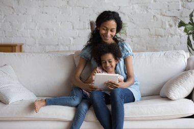 Afrikalı annesini seven küçük kızı dijital tabletle kanepede oturuyor. Yeni eğitim uygulaması kullanan aile, eğlenceli öğrenmenin keyfini çıkarın, genç nesil Alfa ve modern teknoloji kullanımı, geliştirme kavramı