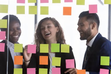 Neşeli çoklu etnik çeşitlilikte iş grubu takım çalışmasını seviyor, cam tahtadaki yapışkan kağıt notları dolduruyor, görevleri tartışıyor, proje planı üzerinde çalışıyor, birlikte program yapıyor, notlar yazıyor, gülüyor, gülüyor.