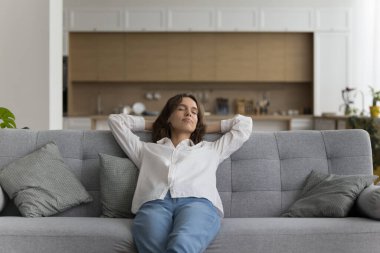 Huzurlu bir kadın rahat bir kanepede dinleniyor, sıcak bir oturma odasında, ellerini başının arkasına koyuyor, modern dairede temiz havanın tadını çıkarıyor. Ev sahibi boş vakitlerini evde dinlenerek geçirir. İklim kontrolü, klima.