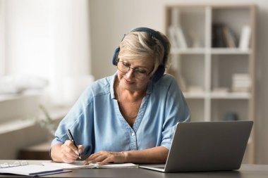 Emeklilik üzerine çalışan, yeni bilgiler edinen, kulaklık takan, deftere yazı yazan, dizüstü bilgisayarlı, online öğrenim oturumuna katılan, sanal toplantı etkinliğine katılan yaşlı bir kadın.