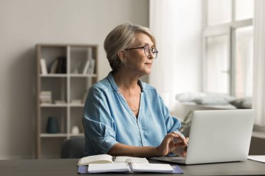 Yaşlı kadın dizüstü bilgisayarını kullanarak masasında oturuyor, dijital aktivitelerle meşgul, pencereden dikkatlice bakıyor, çalışıyor, çalışıyor ya da rahat bir yurt odasında kişisel görevleri yönetiyor. Teknoloji, yaşam tarzı.