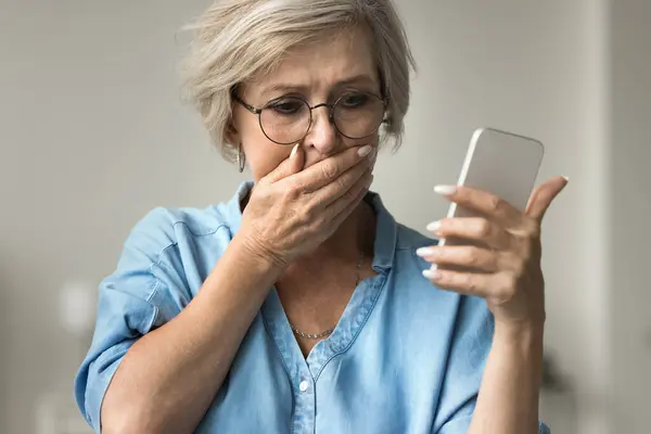 stock image Close up older woman looks at smartphone screen with distressed expression, cover her mouth with hand, read alarming news on phone, feel unhappy, scared. Financial setback, phishing, awful information