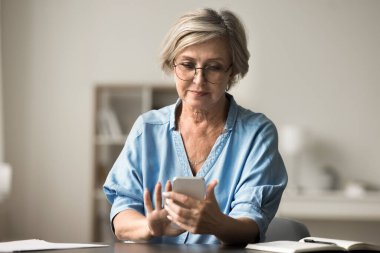 Çekici yaşlı kadın gözlük takıyor akıllı telefonla masaya oturuyor, mesaj gönderiyor, bilgilendirmeleri kontrol ediyor, kablosuz internete bakıyor, iletişim için modern teknolojiyi kullanıyor.