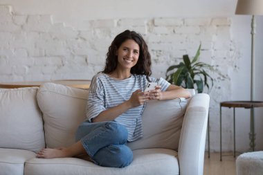 Rahat bir yaşam için modern kablosuz teknoloji kullanımından tatmin olan çekici İspanyol kadın oturma odasındaki rahat koltukta dinleniyor akıllı telefon tebessümü kullanıyor. Uygulama konsepti