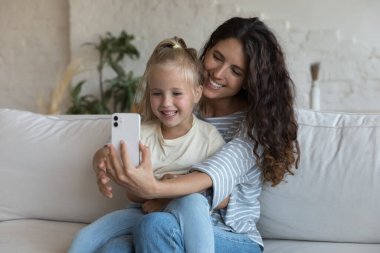 Sevimli genç annesi cep telefonuyla selfie çektirip kanepeye oturur yeni mobil uygulama, komik filtreler ve mutlu görünen yeni vlog videoları kullanarak evde eğlenir. Modern teknoloji konsepti