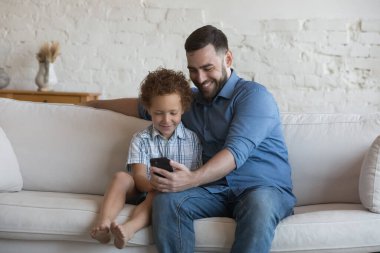 Baba ve anaokulu çocuğu evdeki koltukta oturup cep telefonu kullanıyor. Tatlı kızıl saçlı çocuk. Üvey babası internette eğleniyor. Akıllı telefon, yeni mobil uygulamayla ilgileniyor. Modern teknoloji konsepti