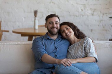 Güzel ev sahipleri, oturma odasındaki rahat koltukta oturup kameraya bakarak hafta sonunun tadını çıkarırlar. Genç aile için banka kredisi, romantik ilişkiler, mutlu evlilik kavramı.
