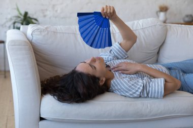 Ateşli Latin bir kadının kanepeye uzanıp el bilgisayarı fanı ile serinlemesinden yorulmuş, tükenmiş görünüyor, kapat. Klima, temiz hava eksikliği, hormonal dengesizlik konsepti.