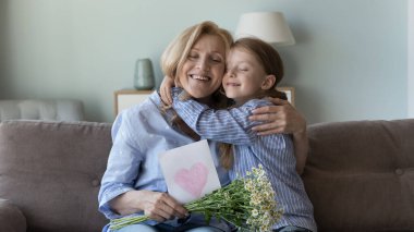 Neşeli, tatlı torun kızı kanepesinde mutlu, minnettar büyükannesine sevgiyle sarılıyor, doğum gününü kutluyor, 8 Mart. Büyükanne elinde kalp, çiçekler ve gülümsemeyle el yapımı bir kart tutuyor.