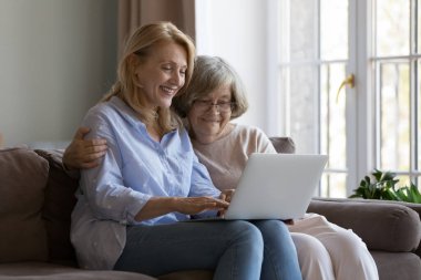 Mutlu yetişkin kız ve yaşlı gri saçlı anne rahat koltukta dizüstü bilgisayarlarını paylaşıyorlar, internet hizmetlerini kullanıyorlar, internet iletişimi için uygulama kullanıyorlar, alışveriş yapıyorlar, sohbet ediyorlar.