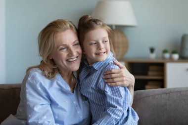 Hayalperest torun kızı, sarışın büyükannesine sevgiyle, sevgiyle ve şefkatle sarılıyor. Büyükanne ve torun kanepede oturuyor, gözlerini kaçırıyor, gülümsüyor. Aile fotoğrafı.