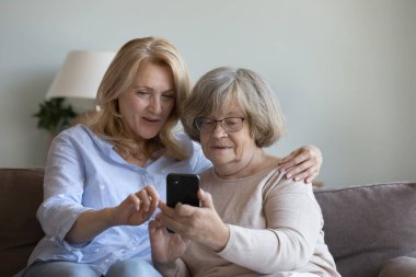 Orta yaşlı bakıcı kadın, yaşlı bayana akıllı telefonda online uygulama kullanmayı öğretiyor. Yetişkin bir kız ve yaşlı bir anne cep telefonuyla konuşuyor, selfie çekiyor, kanepede birlikte dinleniyorlar.