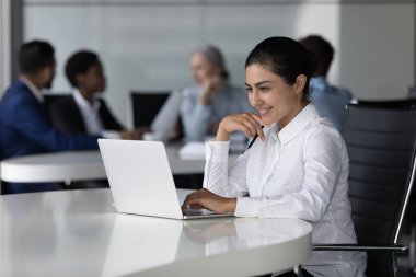 Mutlu güzel Hintli çalışan kız açık ofis çalışma alanındaki dizüstü bilgisayarında başarılı bir online proje üzerinde çalışıyor, ekrana bakıyor, gülümsüyor, iyi haberler alıyor, çalışma masasında oturuyor.