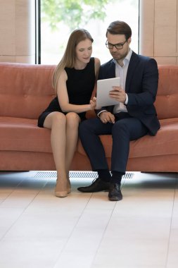 Çalışma molasında tablet paylaşan iki odaklanmış ofis arkadaşı, lobideki koltukta buluşmak, kanepede oturmak, tablet bilgisayardaki online uygulamayı kullanmak, İnternet başlangıç projesini tartışmak. Dikey çekim