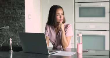 Düşünceli genç serbest yönetici kadın ev mutfağında dizüstü bilgisayar başında çalışıyor, çeneye dokunuyor, başka yere bakıyor, online iş fikirlerini düşünüyor, bilgisayara geçiyor,