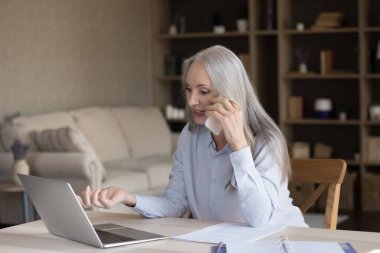 Güzel, orta yaşlı bir kadın akıllı telefonu elinde tutuyor dizüstü bilgisayara bakıyor danışmanıyla online satın alma konuşmalarını tartışıyor, müşteriye yardım ediyor, modern ofiste masa başında oturuyor. İletişim, teknoloji kavramı