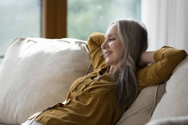 Güzel yaşlı kadın, modern evdeki oturma odasındaki kanepede ellerini başının arkasına koyup tatilin tadını çıkarıyor, temiz havayı soluyor. Refah, konfor yaşam, rahatlama konsepti