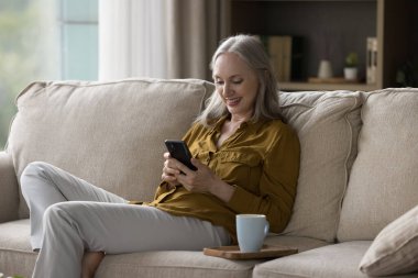 60 'lı yaşlarda rahat bir kadın akıllı telefonuyla kanepede oturuyor. Evde düzen sağlayın, ürünler satın alın perakende elektronik ticari hizmetler kullanın, sohbet edin memnun olun modern teknoloji kullanımı kavramının keyfini çıkarın.
