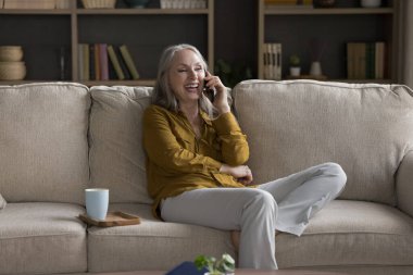 Mutlu, çekici, son sınıf öğrencisi kadın gülüşü çok mutlu. Cep telefonunda keyifli sohbetler. Evde yalnız başına kanepede rahatla. Eski nesil modern kablosuz teknolojiyi, iletişimi, iyi haber kavramını kullanıyor.