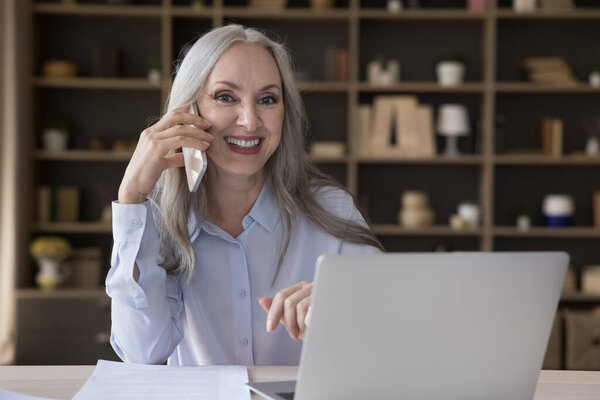 Happy successful mature businesslady talks on smartphone seated at workplace with laptop, having business call looking overjoyed get good news, smiling staring at camera. Modern tech, workflow concept